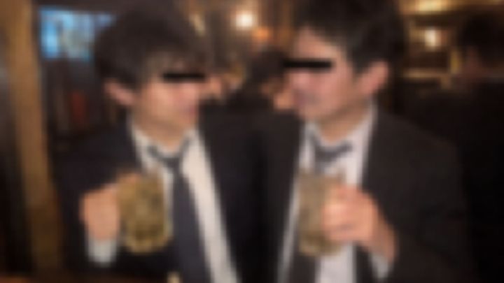立ち飲みの店でジョッキを持って寄り添うスーツの男性2人