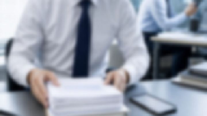 書類を手に机へ座る白いシャツとネクタイ姿の男性