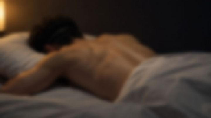 薄暗い部屋で、アイマスクをしたまま裸の背中でベッドにうつ伏せになる男性